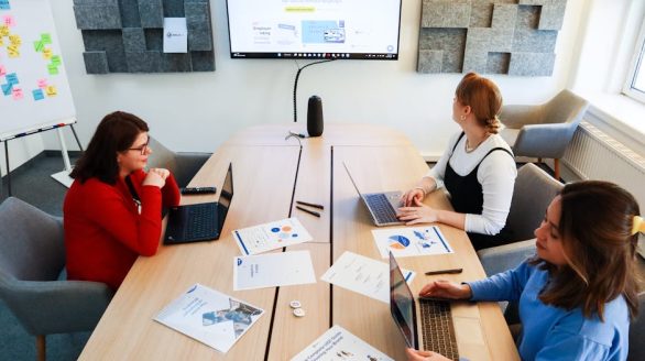 Three women collaborating in a modern office with laptops and presentation materials.